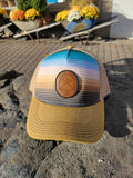 A Joseph, Oregon ball cap with a striped front panel in shades of blue, tan, and gold, featuring a round leather-style patch that reads “Joseph, Oregon – Elev. 4190” with a simple mountain design. The cap has a gold bill, tan mesh back.