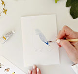 Person painting a bird illustration on white paper with a paintbrush and watercolor.