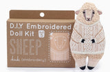 Knitted sheep toy wearing a sweater in front of a DIY embroidered doll kit box.