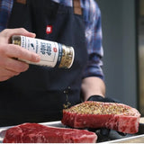 Person seasoning a steak with a spice jar outdoors