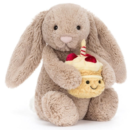 Plush beige bunny with large floppy ears holding a small plush cake with a candle and strawberries.