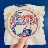 Embroidery hoop with a red and blue mermaid design on a white fabric background.