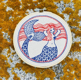 Embroidery hoop with a mermaid design on a textured surface.
