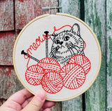 Embroidery hoop with a cat and yarn design held against a wooden background
