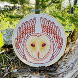 Embroidered owl design in a hoop with a natural background of greenery and rocks.