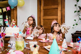 Children at a birthday party blowing party horns around a cake with colorful balloons.