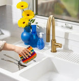a multicolored dishcloth held in someone's hand next to a sink.