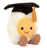 A plush toy boiled egg, wearing a graduation cap with gold tassel. It has a smiling face and brown corduroy legs.